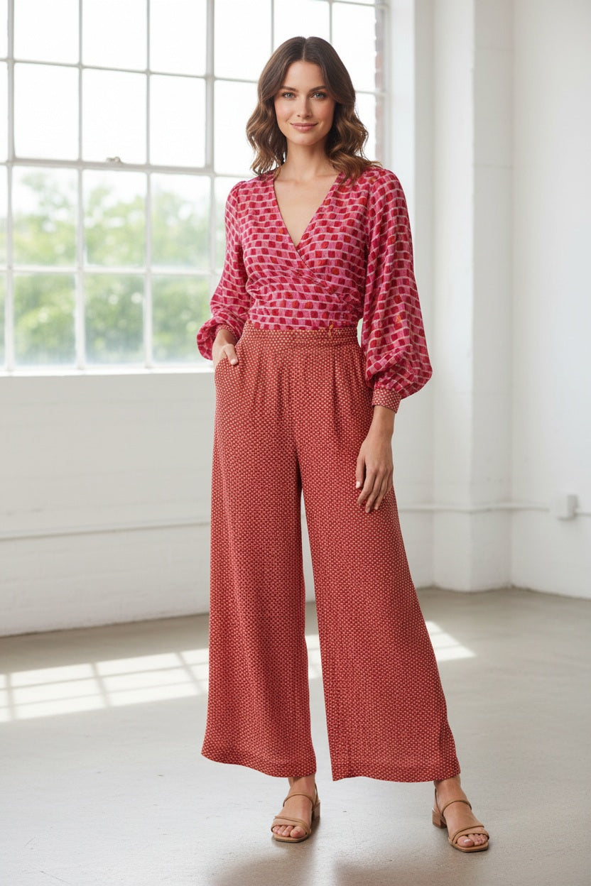 A woman stands against a white background wearing the MIRTO 1956 Mosaic Ramie Trousers, a red patterned blouse, holding a net bag filled with fruit, and white platform shoes.