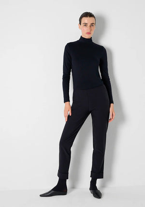 A person stands facing away from the camera against a plain light background, styled in a fitted black turtleneck and KATHARINA HOVMAN - SIMPLE PANTS by KATHARINA HOVMAN, which feature stretch quality for added comfort. Their hair is tied back in a neat, low bun, and black shoes complete the minimalist look.