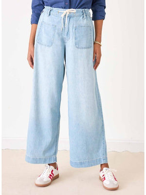 Wearing MERSEA's LONDONBERRY Drawstring Supersoft Denim Pant with wide legs and large front pockets, paired with a light grey top, gold jewelry, and brown sandals, a person poses against a plain light background.