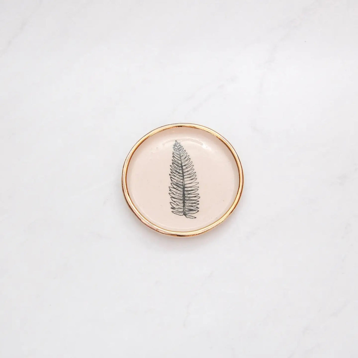 The COMPOST DESIGN - FERN TINY DISH 3&quot; features a black fern leaf on white stoneware, highlighted by a 22k gold rim. Its light, neutral tones and hand-built rustic look beautifully contrast against the white marble surface.