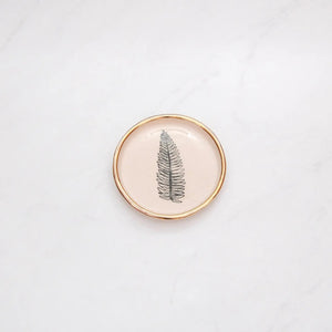 The COMPOST DESIGN - FERN TINY DISH 3" features a black fern leaf on white stoneware, highlighted by a 22k gold rim. Its light, neutral tones and hand-built rustic look beautifully contrast against the white marble surface.