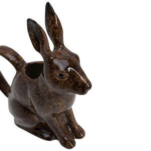 The QUAIL CERAMICS - HARE JUG, LARGE by QUAIL DESIGNS, a delightful ceramic pitcher shaped like a brown rabbit, sits on a wooden surface with white flowers artfully arranged in its open mouth. This charming decorative piece brings whimsy to the tabletop display, while green plants create a softly blurred background.