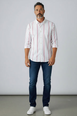 A man in a FRANK & EILEEN Finbar Men's Button Up Shirt in hot pink and blue stripe, blue jeans, and white sneakers walks confidently down a cobblestone street lined with stone buildings and potted plants on a sunny day.