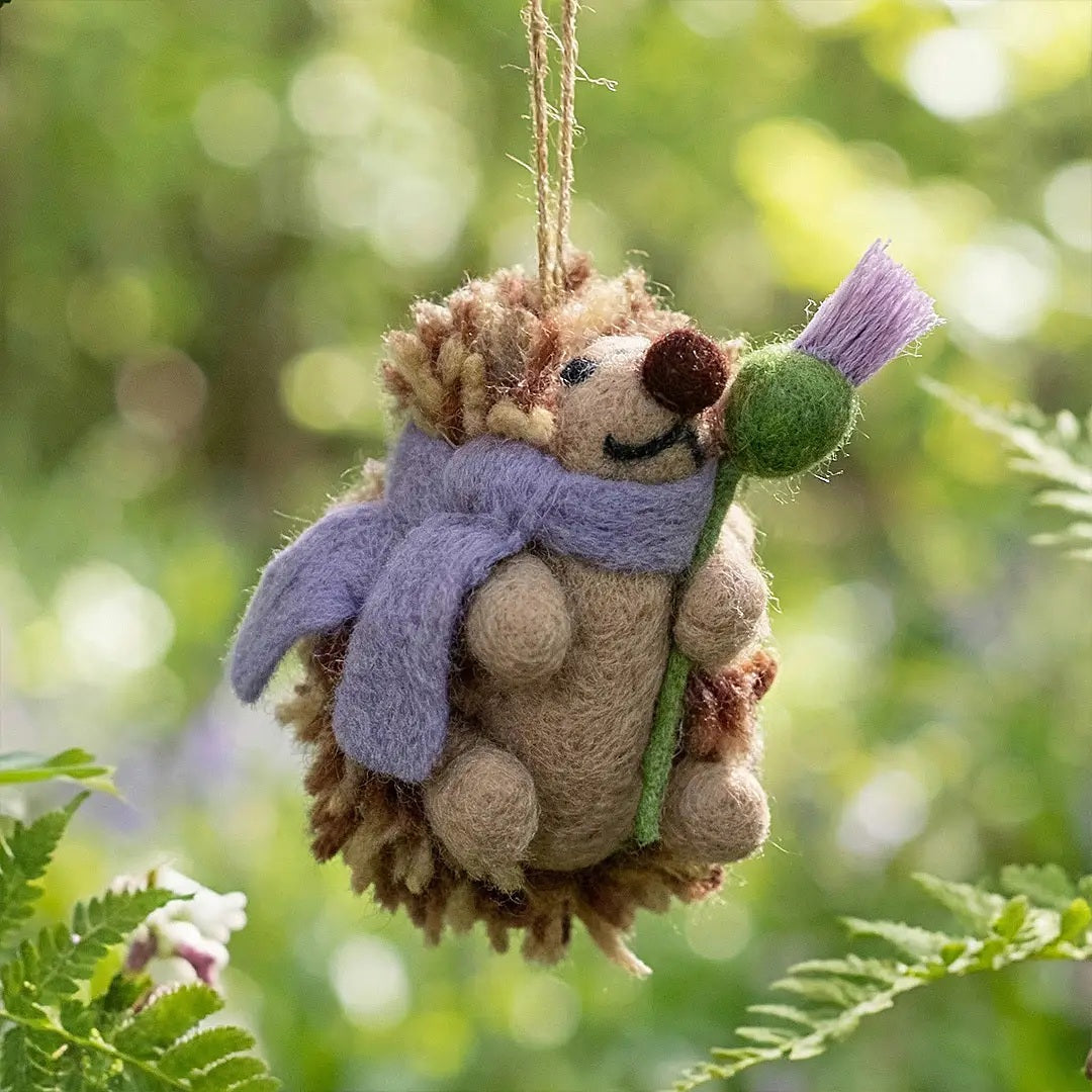A THEA HEDGEHOG WOODLAND ORNAMENT by FELT SO GOOD, designed as a felt Scottish thistle hedgehog wearing a purple scarf and holding a flower, hanging from a twine loop.