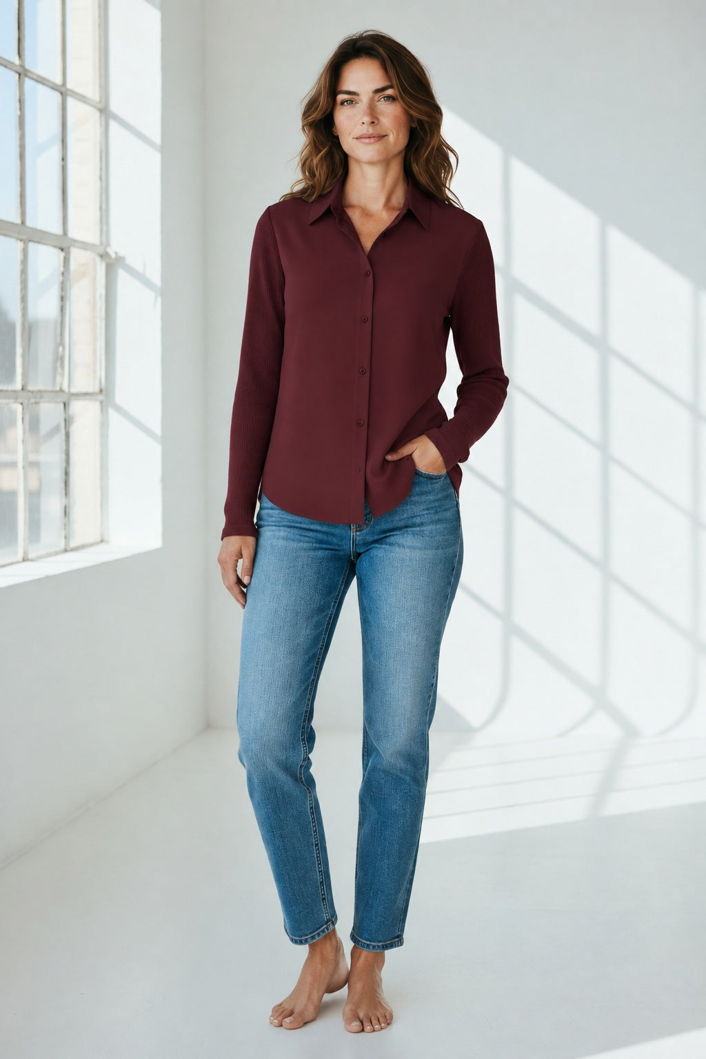 A woman stands barefoot in a bright room, wearing blue jeans and a long-sleeve SEVENTY VENEZIA button-up shirt with modal sleeves in burgundy, one hand tucked into her pocket. Sunlight streams through a large window, casting shadows on the white walls.