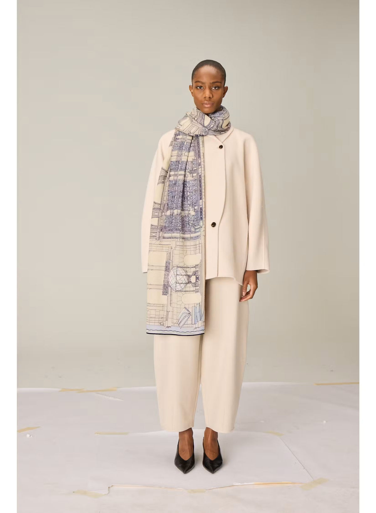 A person wearing a cream-colored outfit stands facing forward, draped in the INOUI EDITIONS VIVIENNE Wool Scarf in Grey—a large, patterned scarf featuring blue and beige tones. Made from 100% wool and crafted in India by INOUI, the scarf measures 100 x 190 cm and is set against a plain, light-colored background.