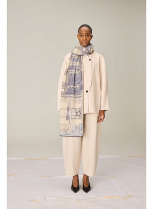 A person wearing a cream-colored outfit stands facing forward, draped in the INOUI EDITIONS VIVIENNE Wool Scarf in Grey—a large, patterned scarf featuring blue and beige tones. Made from 100% wool and crafted in India by INOUI, the scarf measures 100 x 190 cm and is set against a plain, light-colored background.