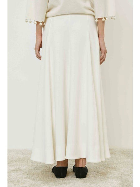 A person stands on a carpeted floor, shown from the back, wearing the MEIMEIJ - SKIRT with a minimalist design and a cream top featuring pearl-detailed sleeves. They wear black shoes against a plain, light background. Brand: MEIMEIJ.