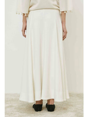 A person stands on a carpeted floor, shown from the back, wearing the MEIMEIJ - SKIRT with a minimalist design and a cream top featuring pearl-detailed sleeves. They wear black shoes against a plain, light background. Brand: MEIMEIJ.