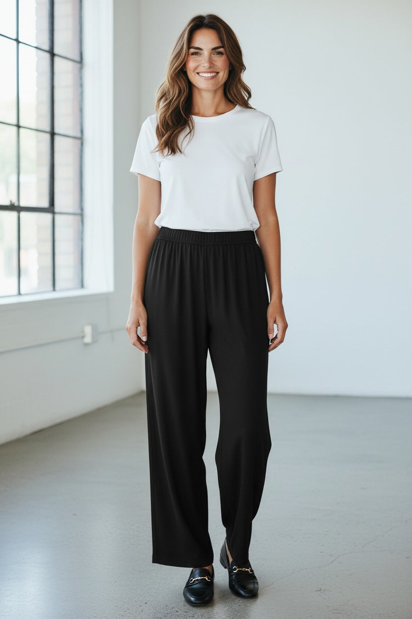 A person wearing MEIMEIJ PULL ON STRAIGHT LEG LONG PANTS in black and a red top, shown from the waist down, stands against a light textured background.