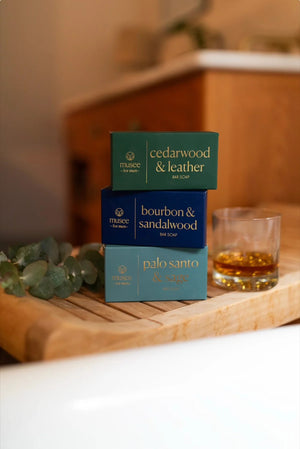 A rectangular white bar of MUSEE - BOURBON & SANDALWOOD BAR SOAP, known for its moisturizing properties, rests partially atop a blue box labeled with the product name and brand “MUSEE,” featuring a circular logo on both the soap and the packaging.