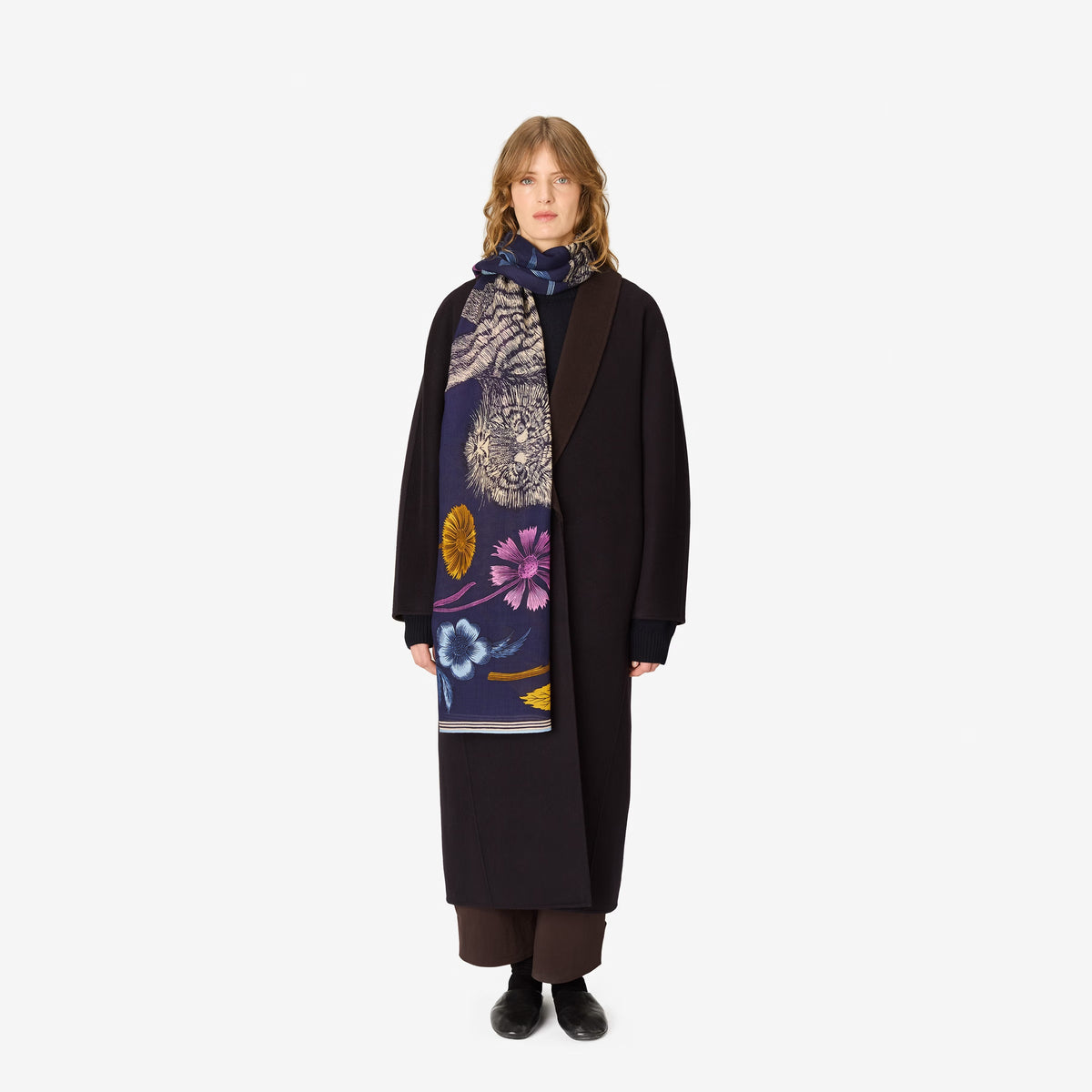 A person stands facing forward against a plain white background, wearing a long dark coat, wide pants, black shoes, and the INOUI EDITIONS - GUSTAV Wool Scarf in Navy by INOUI. An elegant wool rug made in India (80 x 190 cm) is laid beneath their feet.