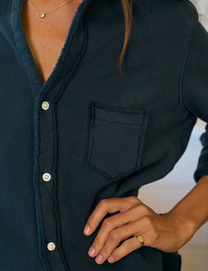 A woman with long brown hair, wearing the FRANK & EILEEN Eileen Relaxed Button Up Sweatshirt in Vintage Navy and light blue jeans, stands in a bright kitchen with white cabinets, a green stove, and a window filled with plants.
