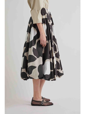 A woman stands against a plain background wearing a beige long-sleeve top, black sandals, and the MEIMEIJ BLACK MOONBEAM SKIRT by MEIMEIJ—a voluminous midi skirt featuring large white and gray abstract shapes.