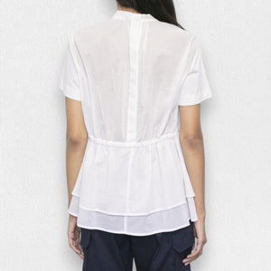 A person is standing against a light textured background wearing the MEIMEIJ - SIDE CINCH TOP, a white short-sleeve, round neck top with side slits and drawstrings. This flared T-shirt features a layered hem and is paired with navy blue pants. Their face is not fully visible.