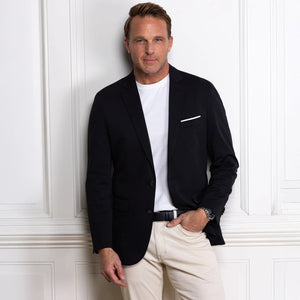 A man in the COLLARS AND CO Maverick Performance Blazer in Stealth Black, layered over a white t-shirt and beige pants, stands by a white paneled wall, looking to the side as he adjusts his blazer.