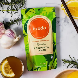 A pouch of BRODO BROTH COMPANY’s BRODO BROTH- TUSCAN SUN ROSEMARY & LEMON BONE BROTH ORGANIC sits on a white surface, surrounded by garlic, lemon, rosemary, and a bowl of broth. This keto-friendly and gluten-free bone broth delivers wholesome goodness to any kitchen.