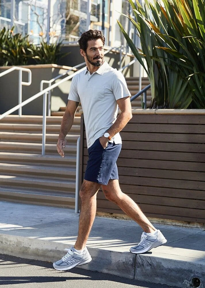A man in a light blue T-shirt, DUER Live Free Journey Shorts in Sapphire, and white sneakers with black details stands with one hand in his pocket against a plain light background.