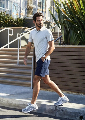 A man in a light blue T-shirt, DUER Live Free Journey Shorts in Sapphire, and white sneakers with black details stands with one hand in his pocket against a plain light background.
