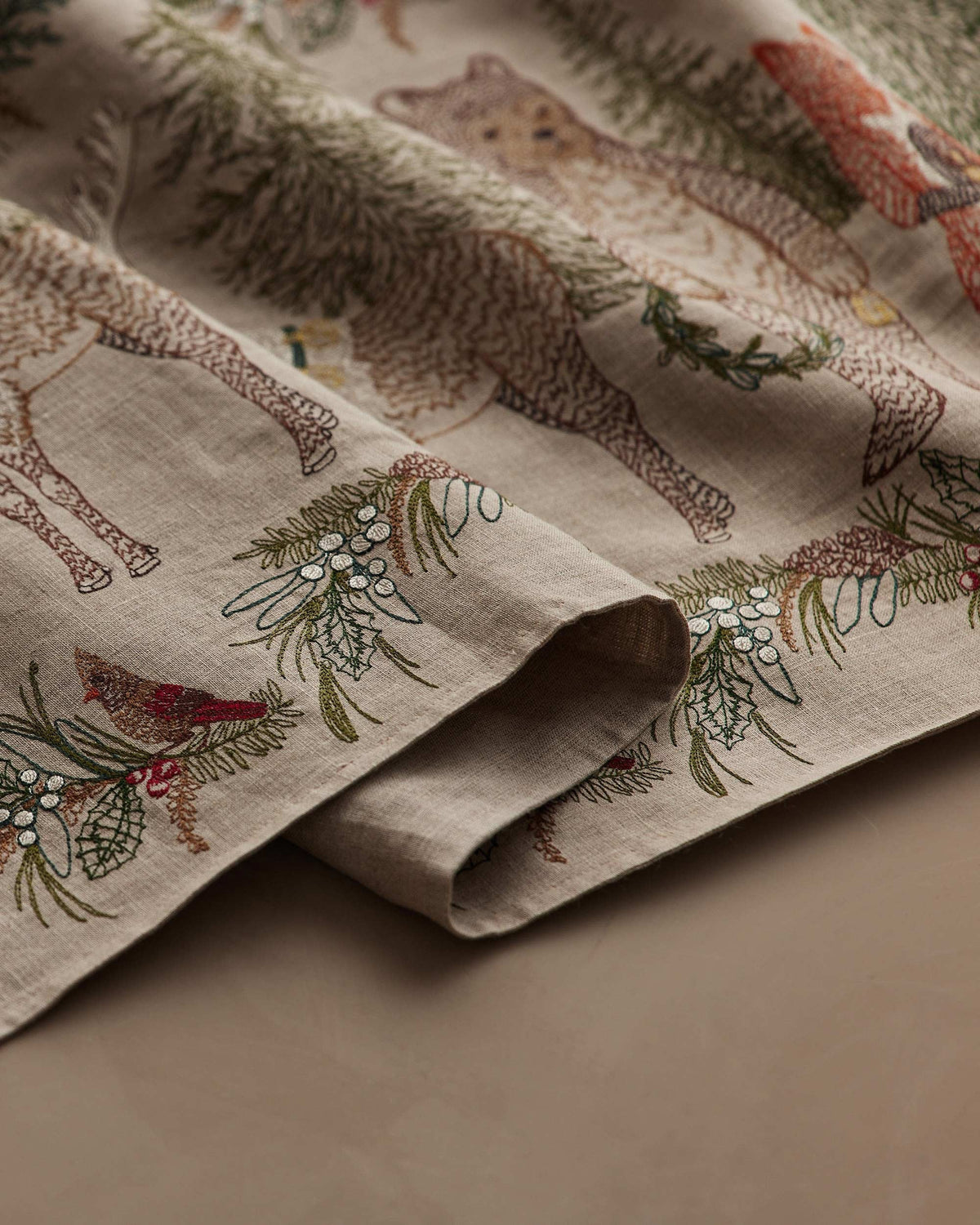 A CHRISTMAS CHEER EMBROIDERED LINEN TABLE RUNNER featuring woodland animals in winter attire and Christmas trees, draped over a wooden dining table set with plates, glassware, and candlesticks.