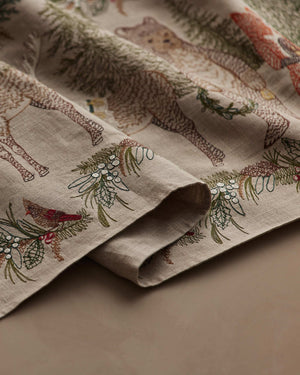 A CHRISTMAS CHEER EMBROIDERED LINEN TABLE RUNNER featuring woodland animals in winter attire and Christmas trees, draped over a wooden dining table set with plates, glassware, and candlesticks.