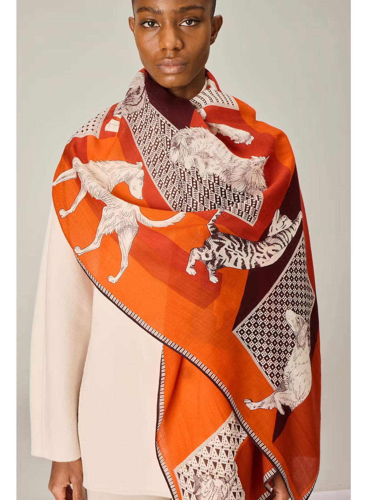 A person dressed in a cream outfit models the INOUI EDITIONS - LES CHERIS WOOL SCARF IN RED by INOUI—a 100% wool accessory made in India, featuring bold orange, red, and burgundy tones adorned with large white cat illustrations and geometric patterns—set against a plain background.