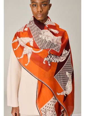 A person dressed in a cream outfit models the INOUI EDITIONS - LES CHERIS WOOL SCARF IN RED by INOUI—a 100% wool accessory made in India, featuring bold orange, red, and burgundy tones adorned with large white cat illustrations and geometric patterns—set against a plain background.
