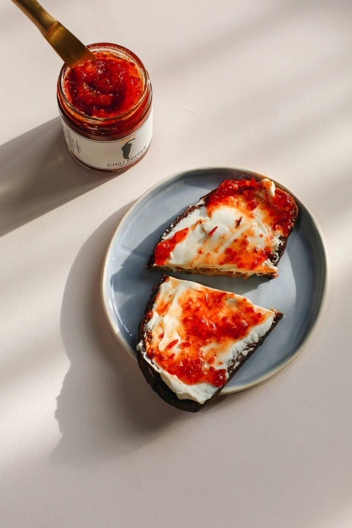 A glass jar of BRINS - CHILI PEPER SPREAD by BRINS, made with garlic, ginger, lime, red jalapeños, and finished with a splash of apple cider vinegar for a tangy depth. The vibrant red-orange spicy chili jam comes in a jar topped with a black lid; the label showcases an illustration of a black chili pepper alongside product details.