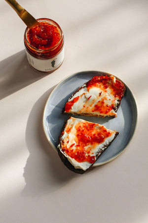 A glass jar of BRINS - CHILI PEPER SPREAD by BRINS, made with garlic, ginger, lime, red jalapeños, and finished with a splash of apple cider vinegar for a tangy depth. The vibrant red-orange spicy chili jam comes in a jar topped with a black lid; the label showcases an illustration of a black chili pepper alongside product details.