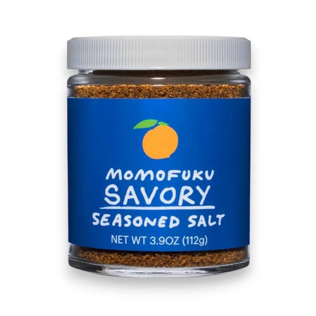 A clear jar with a white lid holds brown MOMOFUKU - SAVORY SALT, featuring a blue label with an orange fruit graphic and "Momofuku Savory Seasoned Salt" text. Net weight: 3.9 oz (112g). Made with premium kosher salt. Brand: MOMOFUKU.
