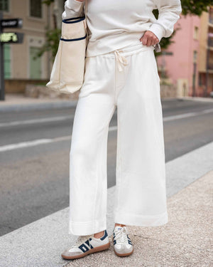 A person stands on a sidewalk in front of urban buildings and a street, wearing a white sweatshirt paired with the MERSEA - FRENCHIE WIDE LEG PANT IN PERIWINKLE from MERSEA. The relaxed fit French terry pants feature a wide-leg silhouette and are styled with sporty beige-and-blue sneakers and a cream tote bag with dark trim.