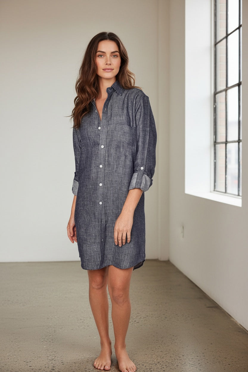 A person stands against a plain light background, smiling, in the EMILY PHILLIPS - THE LINEN DENIM MINI SHIRTDRESS from EMILY PHILLIPS—a long-sleeve, button-down dress made from sustainable linen with a dark chambray look—styled with rolled-up sleeves and white sneakers.