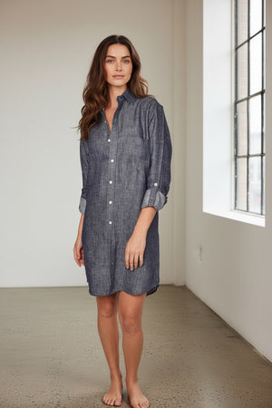A person stands against a plain light background, smiling, in the EMILY PHILLIPS - THE LINEN DENIM MINI SHIRTDRESS from EMILY PHILLIPS—a long-sleeve, button-down dress made from sustainable linen with a dark chambray look—styled with rolled-up sleeves and white sneakers.