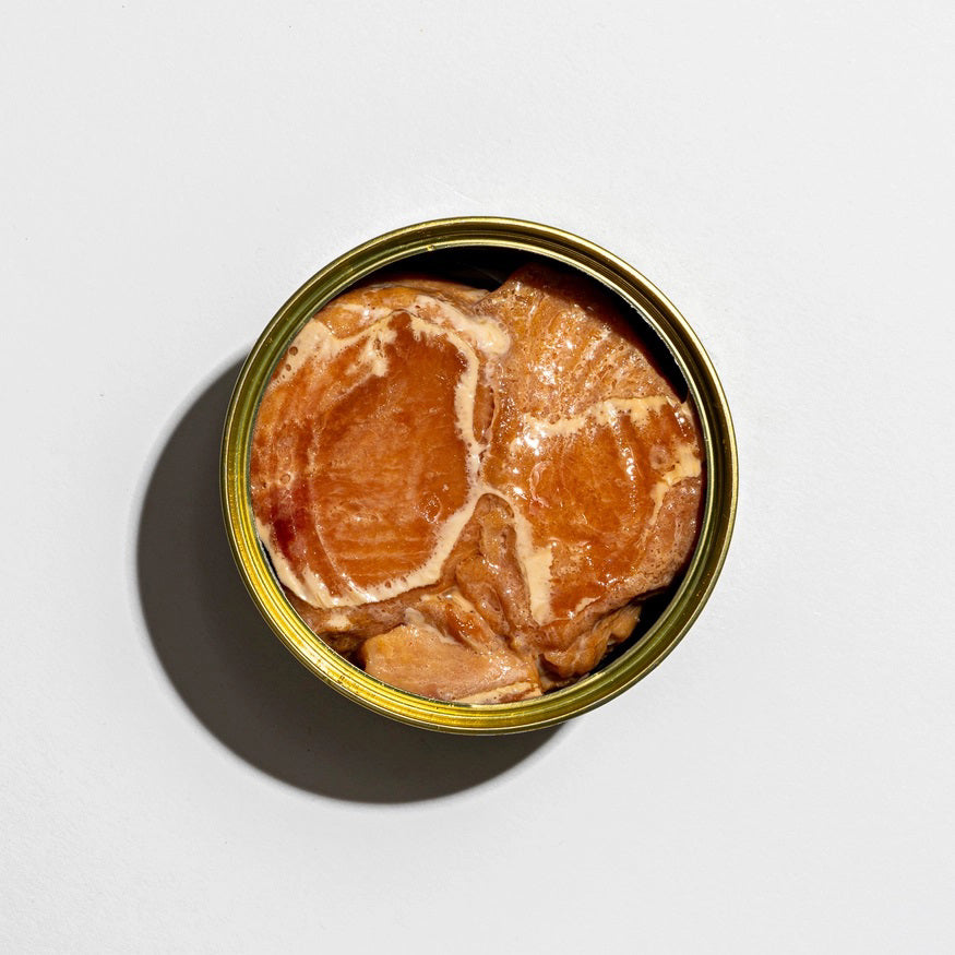 A vibrant square package of FISHWIFE - WILD CAUGHT SMOKED ALBACORE TUNA set against a white backdrop. The packaging showcases an illustrated fish wearing a hat, surrounded by text and decorative accents. Made from MSC-certified albacore tuna sourced from British Columbia and smoked in small batches, the main colors featured are pink, yellow, blue, and green.