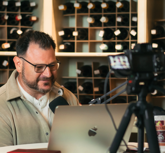 A man with glasses sits at a desk with a laptop and microphone, speaking in front of a camera. Wine bottles are organized in cubbies on the wall behind him.