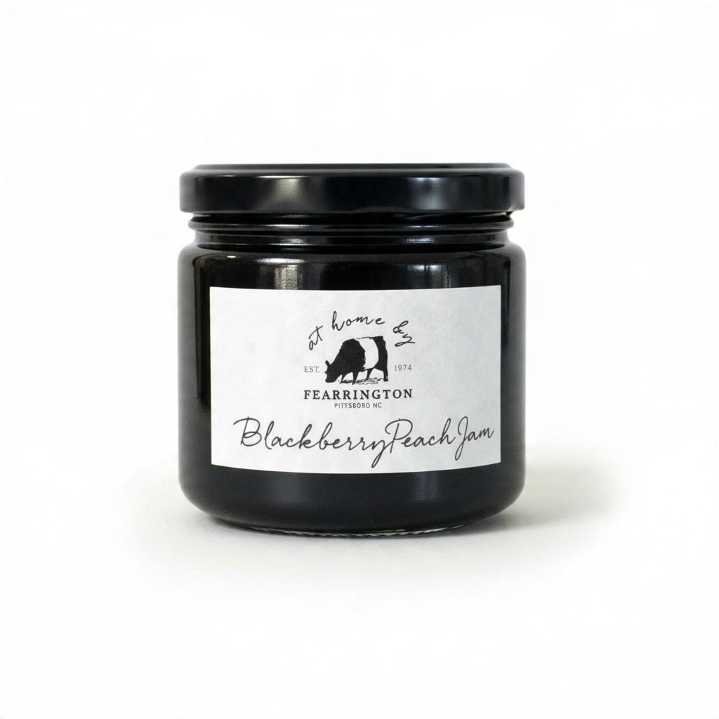 A glass jar of FEARRINGTON SIGNATURE COLLECTION - FEARRINGTON HOUSEMADE BLACKBERRY PEACH JAM with a black lid and a white label. The label lists ingredients: blackberries, peaches, lemon, cinnamon, nutmeg, sugar, pectin. It also mentions: "Handmade with care in our kitchens by Fearrington chefs." Perfect as a hostess gift or for breakfast jams. May contain allergens.