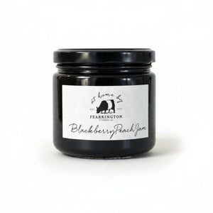 A glass jar of FEARRINGTON SIGNATURE COLLECTION - FEARRINGTON HOUSEMADE BLACKBERRY PEACH JAM with a black lid and a white label. The label lists ingredients: blackberries, peaches, lemon, cinnamon, nutmeg, sugar, pectin. It also mentions: "Handmade with care in our kitchens by Fearrington chefs." Perfect as a hostess gift or for breakfast jams. May contain allergens.