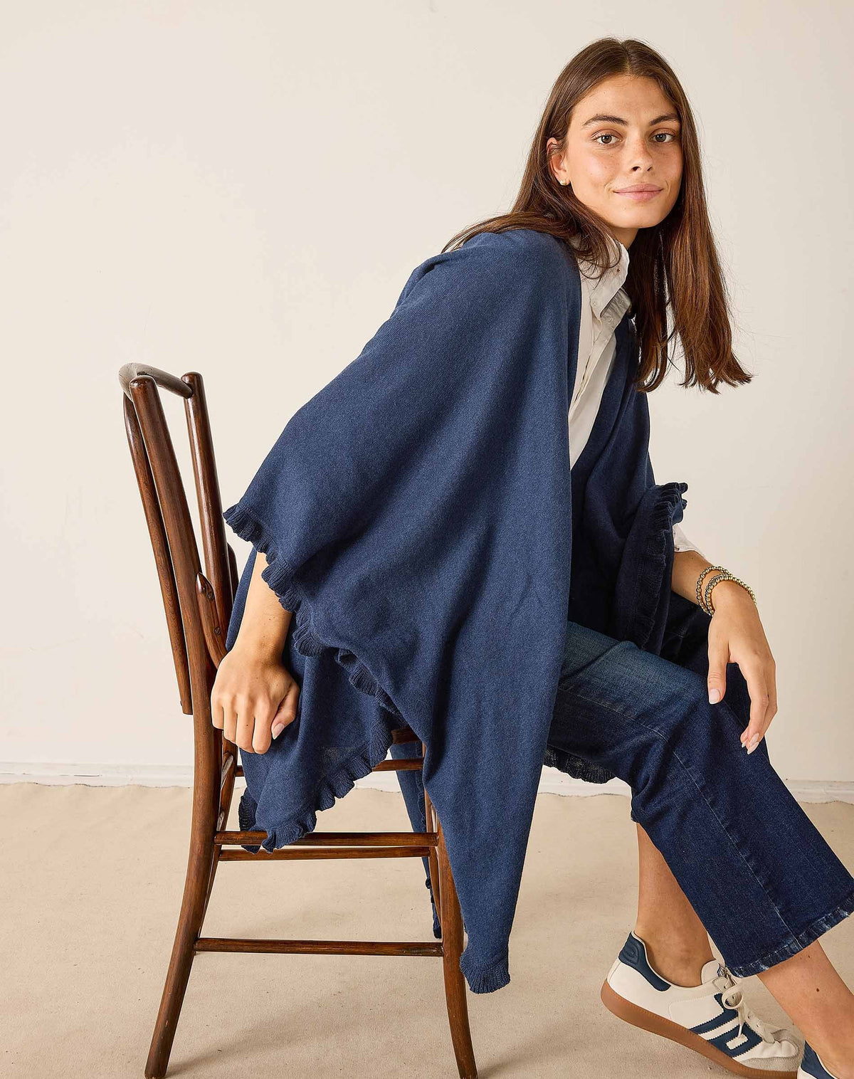 A woman stands sideways against a plain background, dressed in a white shirt and blue jeans. She styles her look with the MERSEA - CHARLESTON RUFFLE WRAP IN CHAMBRAY by MERSEA, worn as a loose navy shawl. Her outfit is completed with white sneakers featuring black stripes, while her long brown hair frames her face as she looks at the camera.