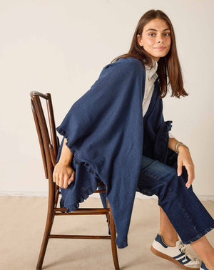 A woman stands sideways against a plain background, dressed in a white shirt and blue jeans. She styles her look with the MERSEA - CHARLESTON RUFFLE WRAP IN CHAMBRAY by MERSEA, worn as a loose navy shawl. Her outfit is completed with white sneakers featuring black stripes, while her long brown hair frames her face as she looks at the camera.