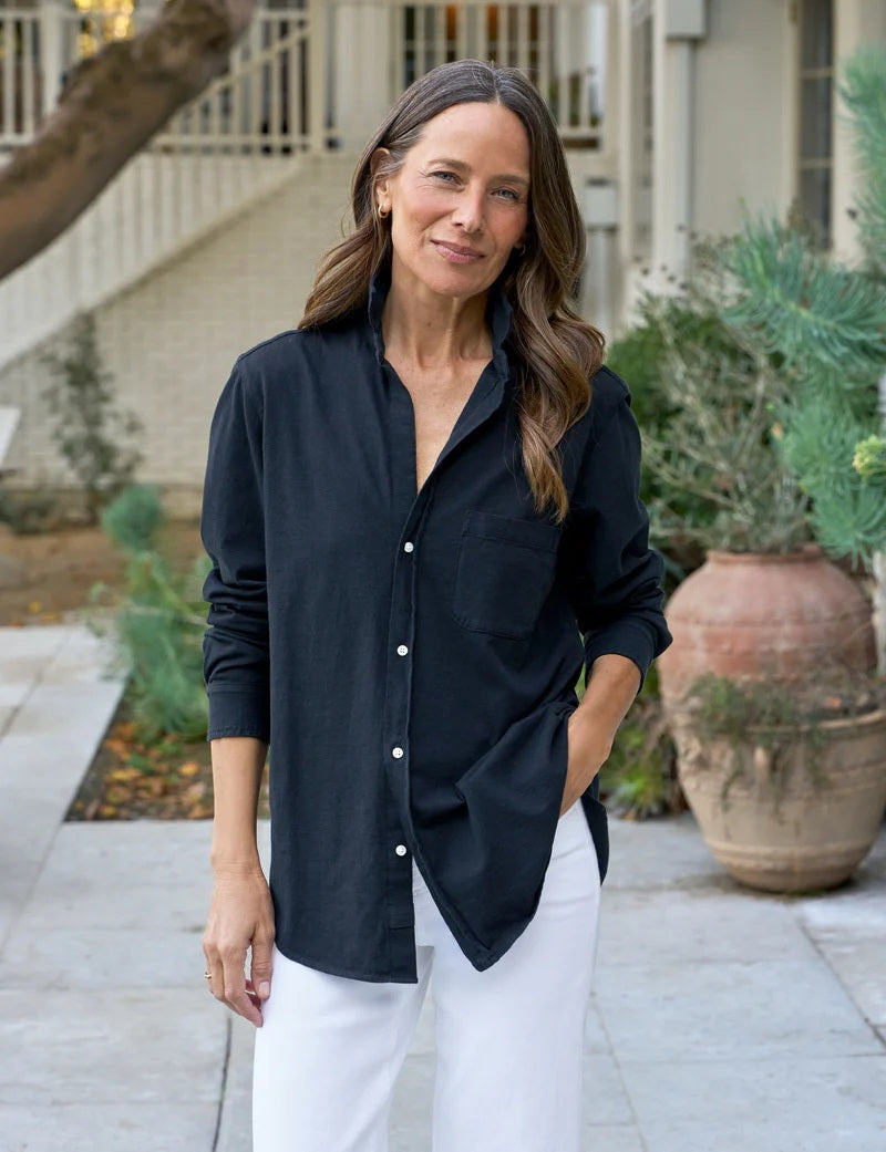 A woman with straight brown hair smiles on a patio, wearing the FRANK &amp; EILEEN Waverly NYC Button Up Shirt in Vintage Jersey British Royal Navy, paired with white pants. Plants and large pots decorate the background.