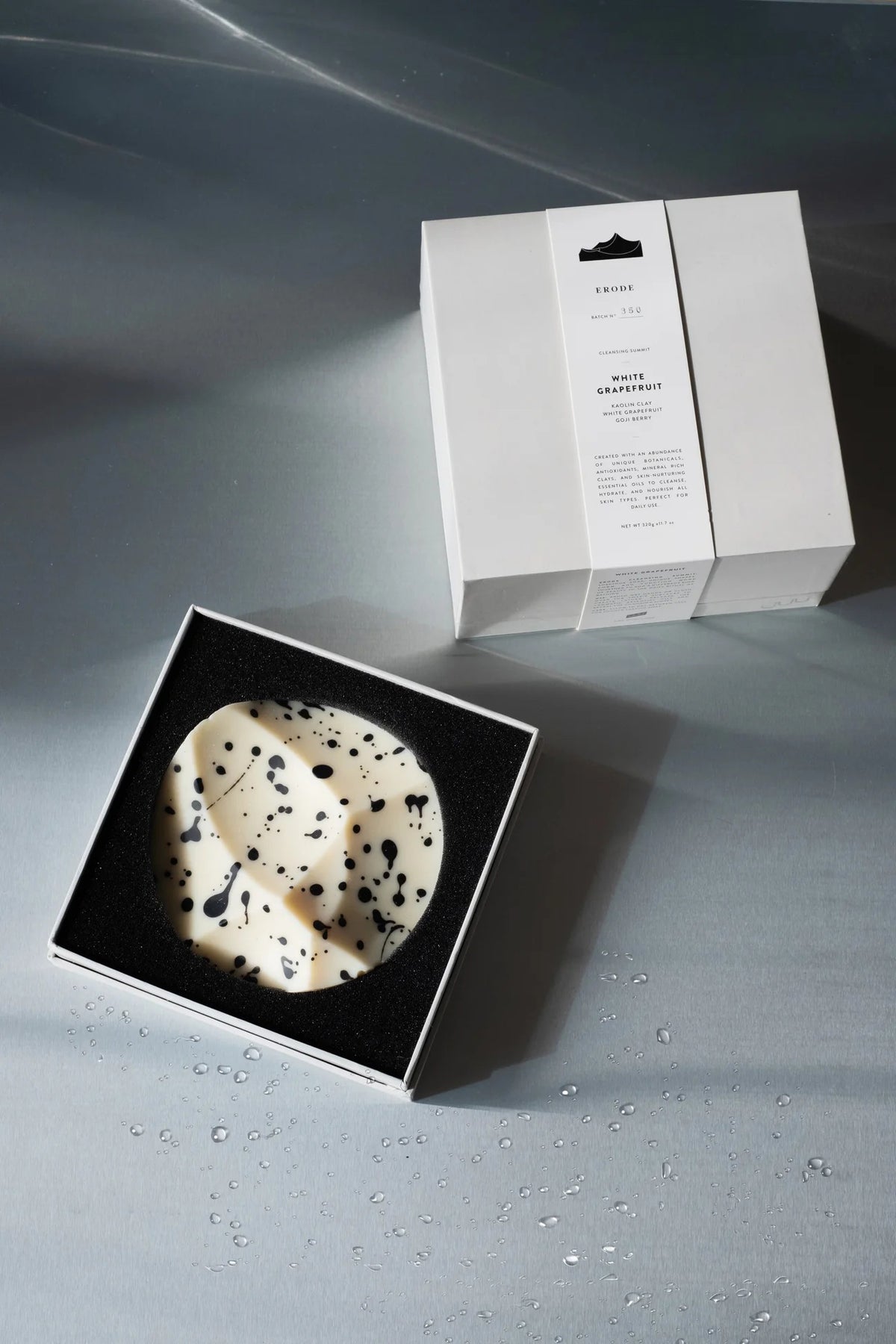 The UME STUDIO UME - WHITE GRAPEFRUIT ERODE SOAP, a round white handmade bar with black speckles, rests in a black foam-lined box beside its white packaging, both displayed on a silver surface with water droplets.