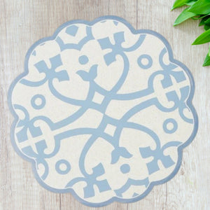 The ETUHOME Round Scalloped Placemat in Denim, featuring a blue and white geometric floral pattern, is displayed on a light wooden surface with green plant leaves visible in the upper right corner.