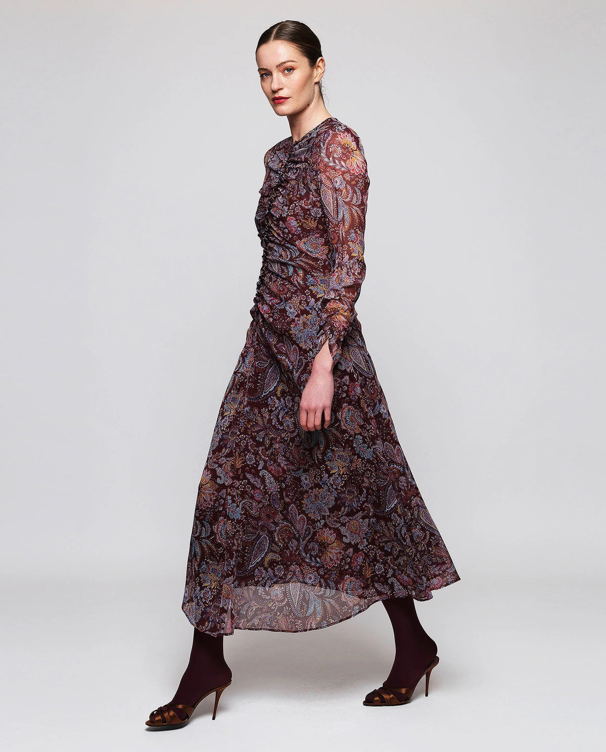 A woman stands facing away, wearing the MIRTO 1956 Burgundy Draped Paisley Print Dress—a long sleeve midi dress featuring an elegant paisley pattern—paired with burgundy tights and black heels. Her hair is styled in a neat bun as she poses against a plain white background.