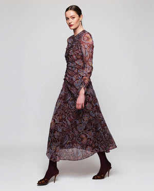 A woman stands facing away, wearing the MIRTO 1956 Burgundy Draped Paisley Print Dress—a long sleeve midi dress featuring an elegant paisley pattern—paired with burgundy tights and black heels. Her hair is styled in a neat bun as she poses against a plain white background.