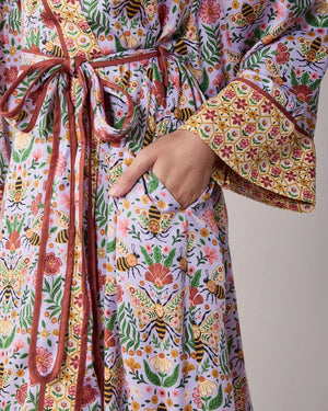A woman wears a PRINTFRESH - BEE TAPESTRY ECO SATIN ROBE IN LAVENDER, featuring a lavender floral-print with a repeating pattern of bees, flowers, and vines with a contrasting yellow and red patterned trim.