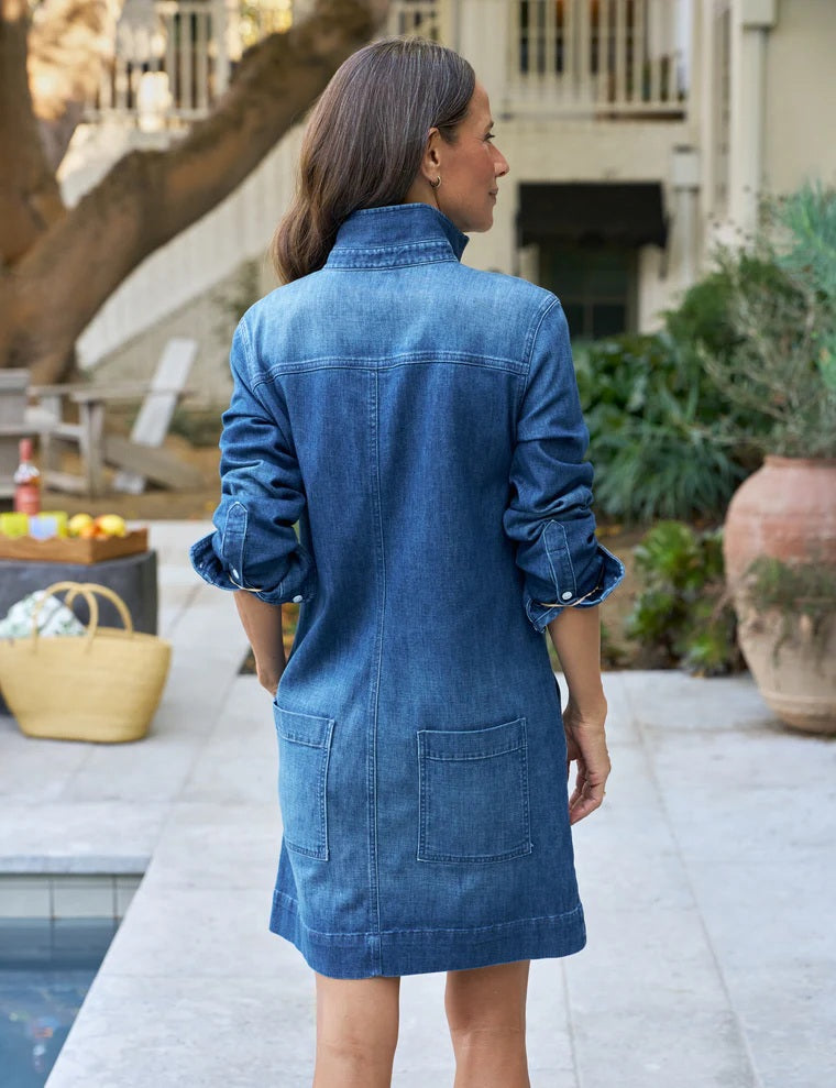A person wearing the FRANK & EILEEN Southern Ireland Utility Dress in Italian Dream Light Denim 1984 Wash, featuring long sleeves, large front pockets, and white buttons, stands outdoors with one hand in their pocket. Their face is not visible. The dress’s classic style subtly nods to practical Irish design.