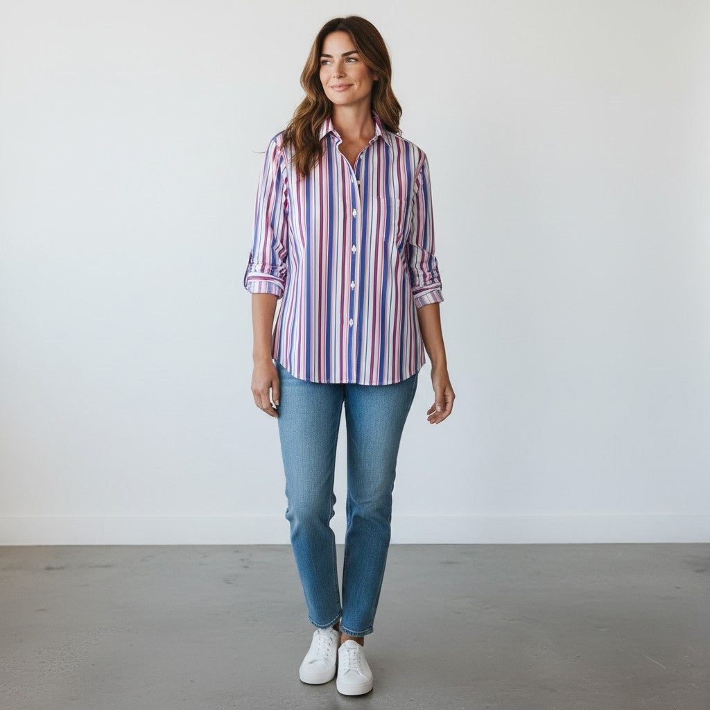 A woman sits outdoors on a wicker chair, smiling at the camera, wearing the FRANK & EILEEN EILEEN Relaxed Button Up in Italian Cotton Multicolor Stripe paired with jeans. Sunlight filters over green plants and flowers in the background.