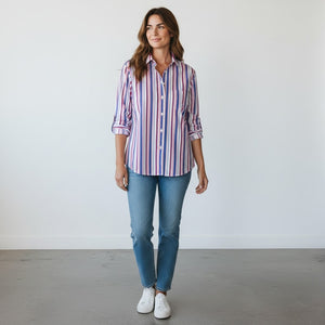 A woman sits outdoors on a wicker chair, smiling at the camera, wearing the FRANK & EILEEN EILEEN Relaxed Button Up in Italian Cotton Multicolor Stripe paired with jeans. Sunlight filters over green plants and flowers in the background.