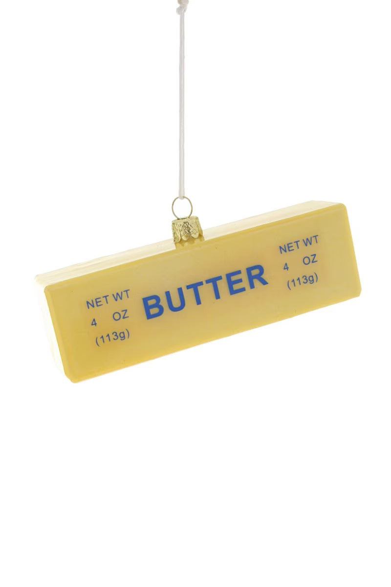 The CODY FOSTER STICK OF BUTTER ORNAMENT is a rectangular glass decoration with &quot;BUTTER&quot; in blue and a 4 oz (113g) net weight, hanging by a string against a white background.