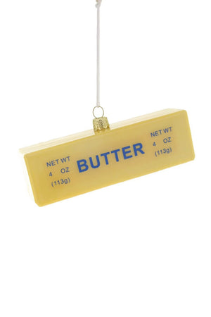 The CODY FOSTER STICK OF BUTTER ORNAMENT is a rectangular glass decoration with "BUTTER" in blue and a 4 oz (113g) net weight, hanging by a string against a white background.